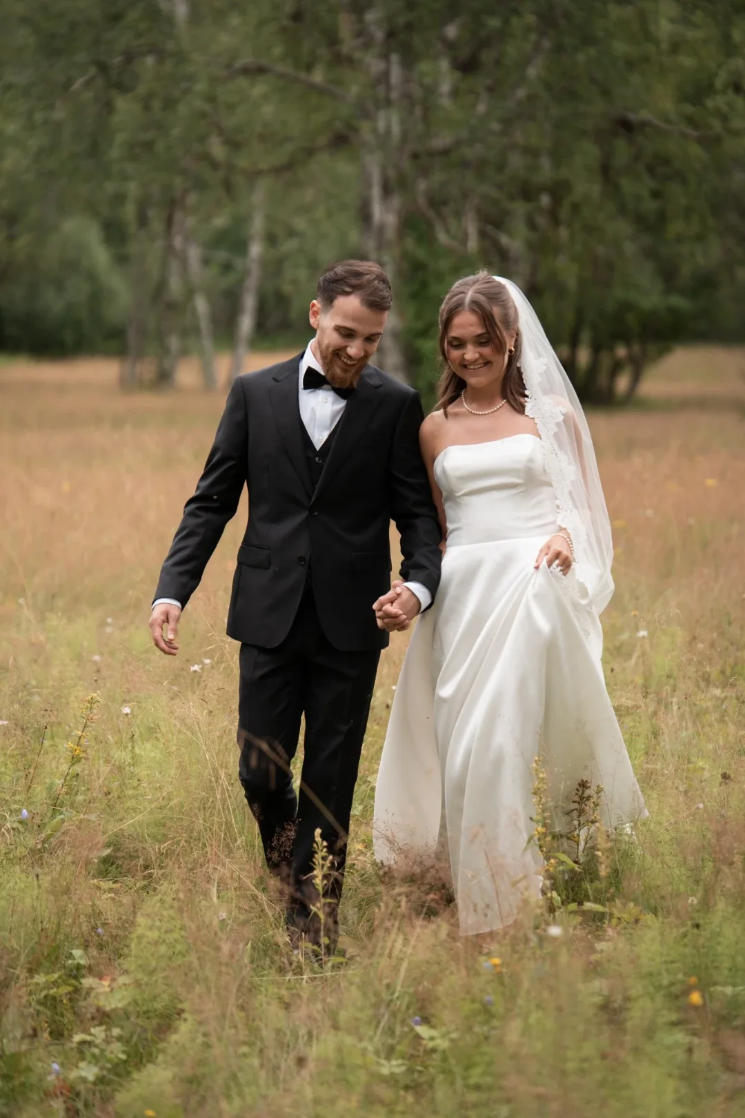 A smiling bride and groom walking hand in hand through a summer meadow