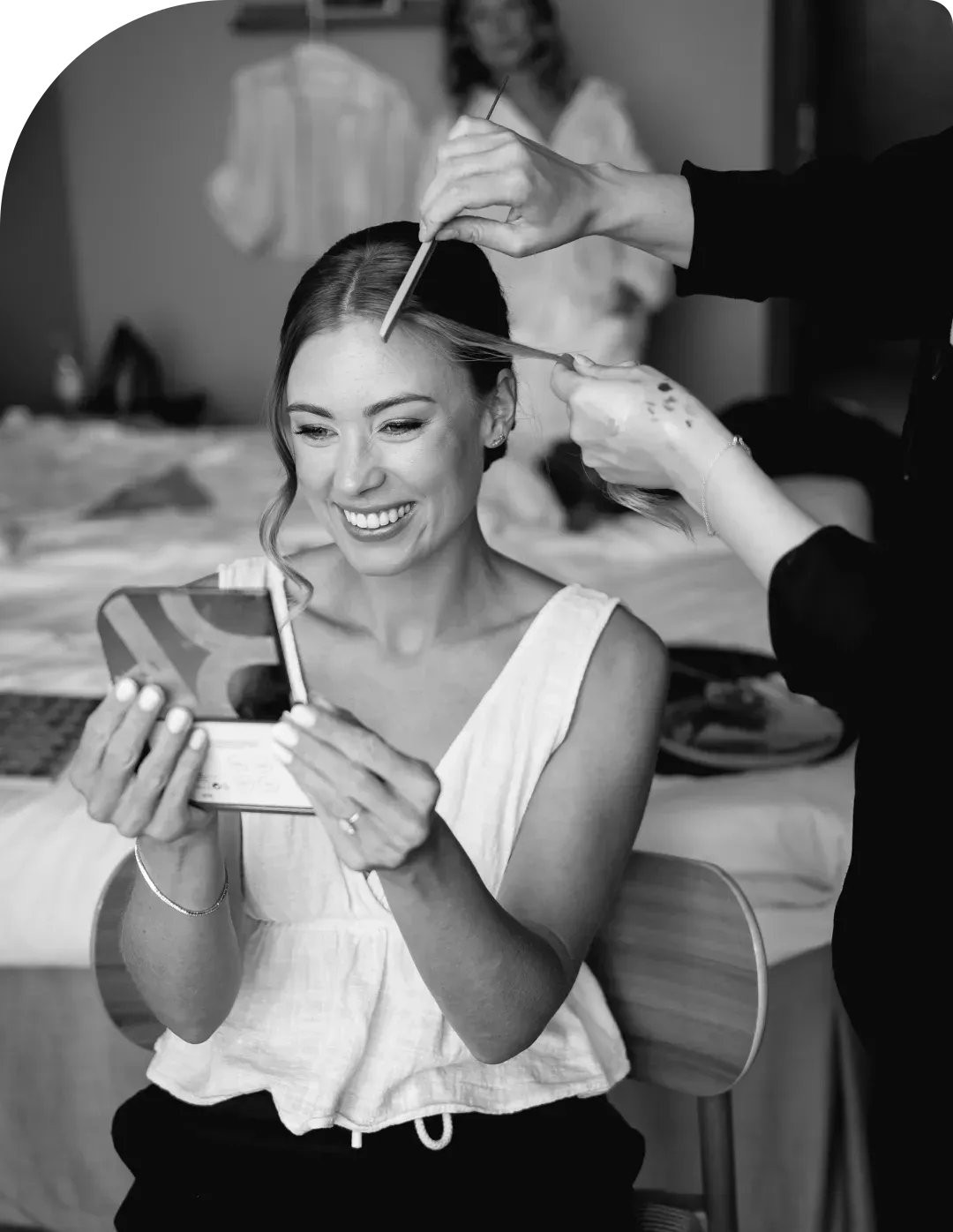 A bride laughing with a mirror in her hand while her makeup is done, black and white